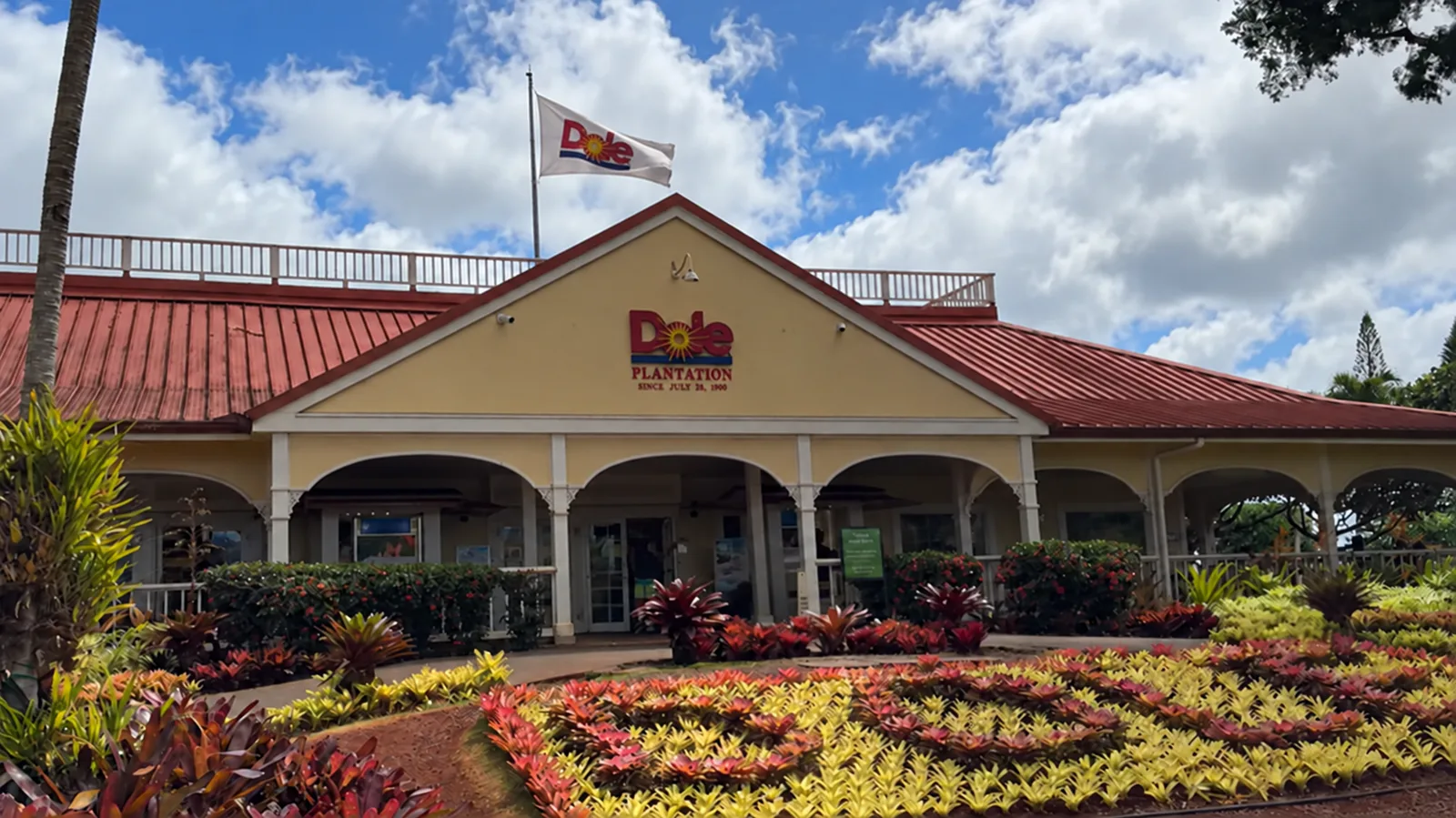 赤い屋根とクリーム色の壁の建物。正面に「Dole PLANTATION」の看板と、屋根の上にドールの旗。手前の花壇には赤と黄色のブロメリアが並ぶ