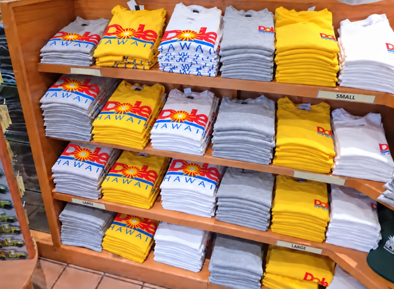 木の棚に積まれた白・黄色・グレーのTシャツ。胸に「Dole HAWAII」のロゴが大きく入っている