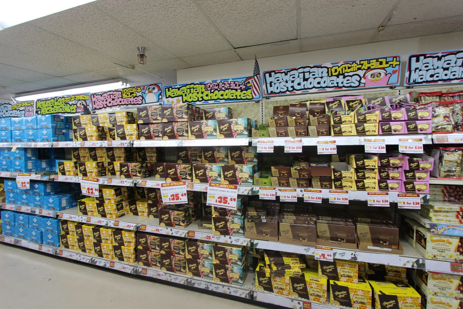「Hawaiian Host Chocolates」の看板の下に、金色や茶色のハワイアンホーストのチョコの箱が棚いっぱいに並ぶ