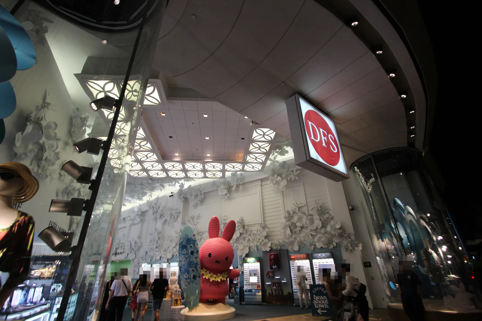 夜のカラカウア通り側から見た、赤い丸のDFSの看板が掲げられた免税店の入り口。白い花の装飾の前に、レイをかけたピンクのうさぎの大きな置物とサーフボードが飾られている
