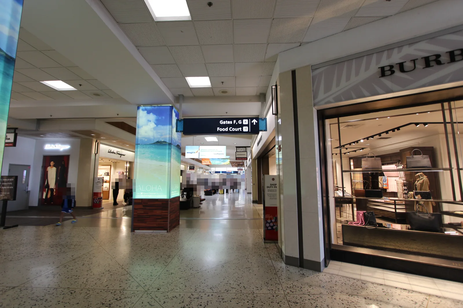 ホノルル空港の出発エリアの通路。右手にブランドのブティック、左手にも免税店が並び、天井から「Gates F, G」「Food Court」の案内板が下がっている