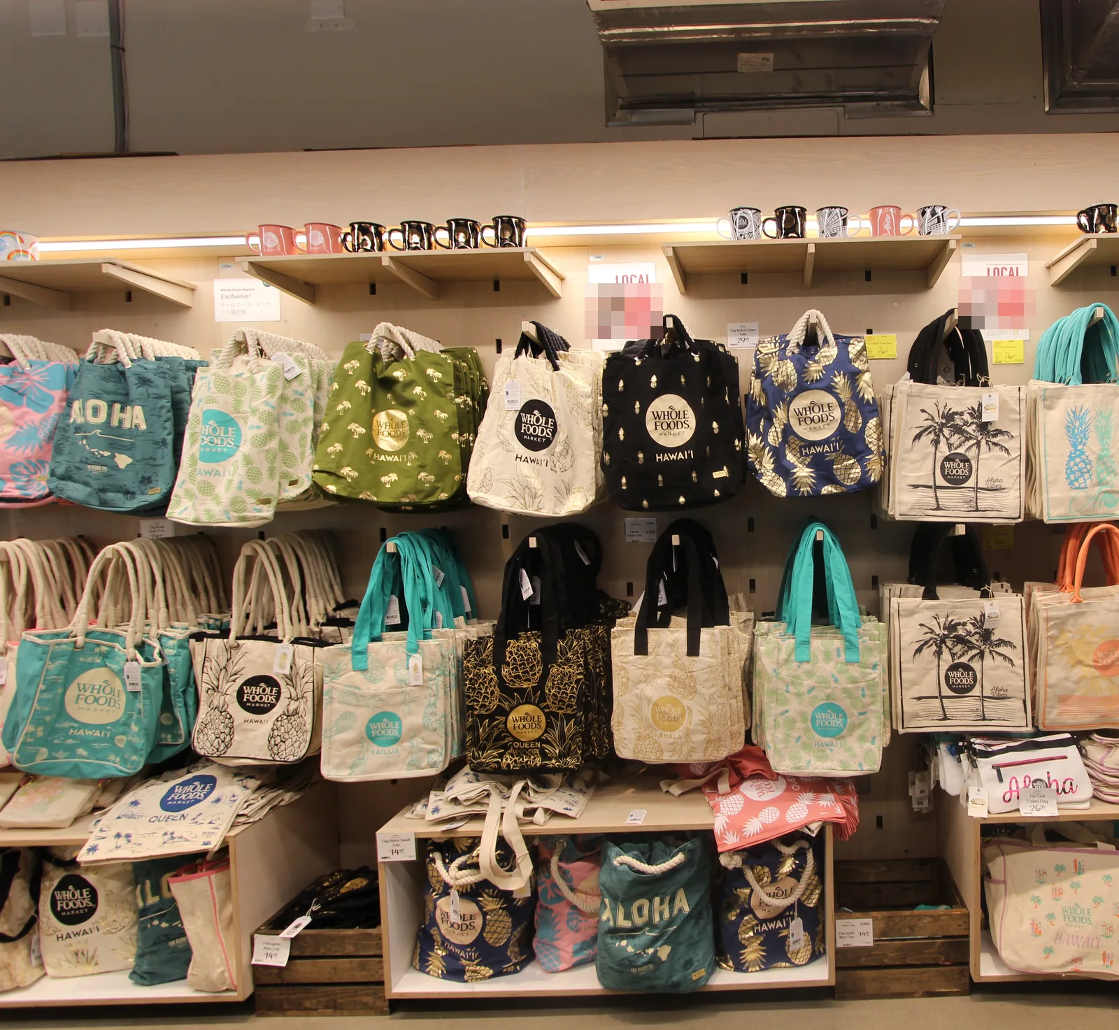 ホールフーズのハワイ限定トートバッグの売り場。HAWAI'I や QUEEN・KAILUA・KAHALA の店舗名、ALOHA の文字、パイナップル柄などのバッグが棚に並ぶ