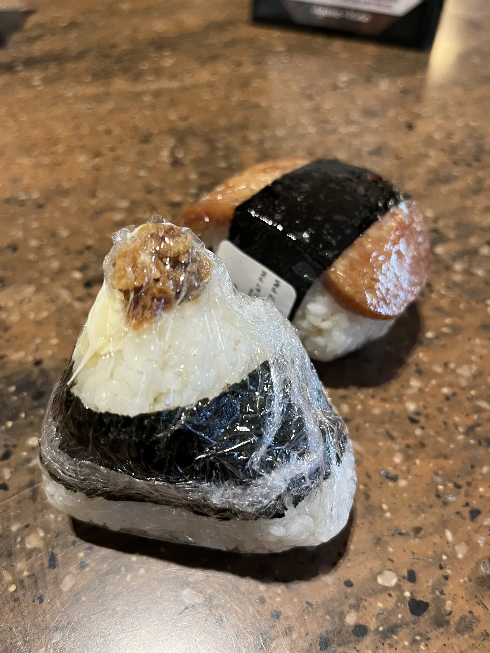 ラップに包まれた三角のおにぎりと、奥に照り焼きのスパムむすびがテーブルに並んでいる
