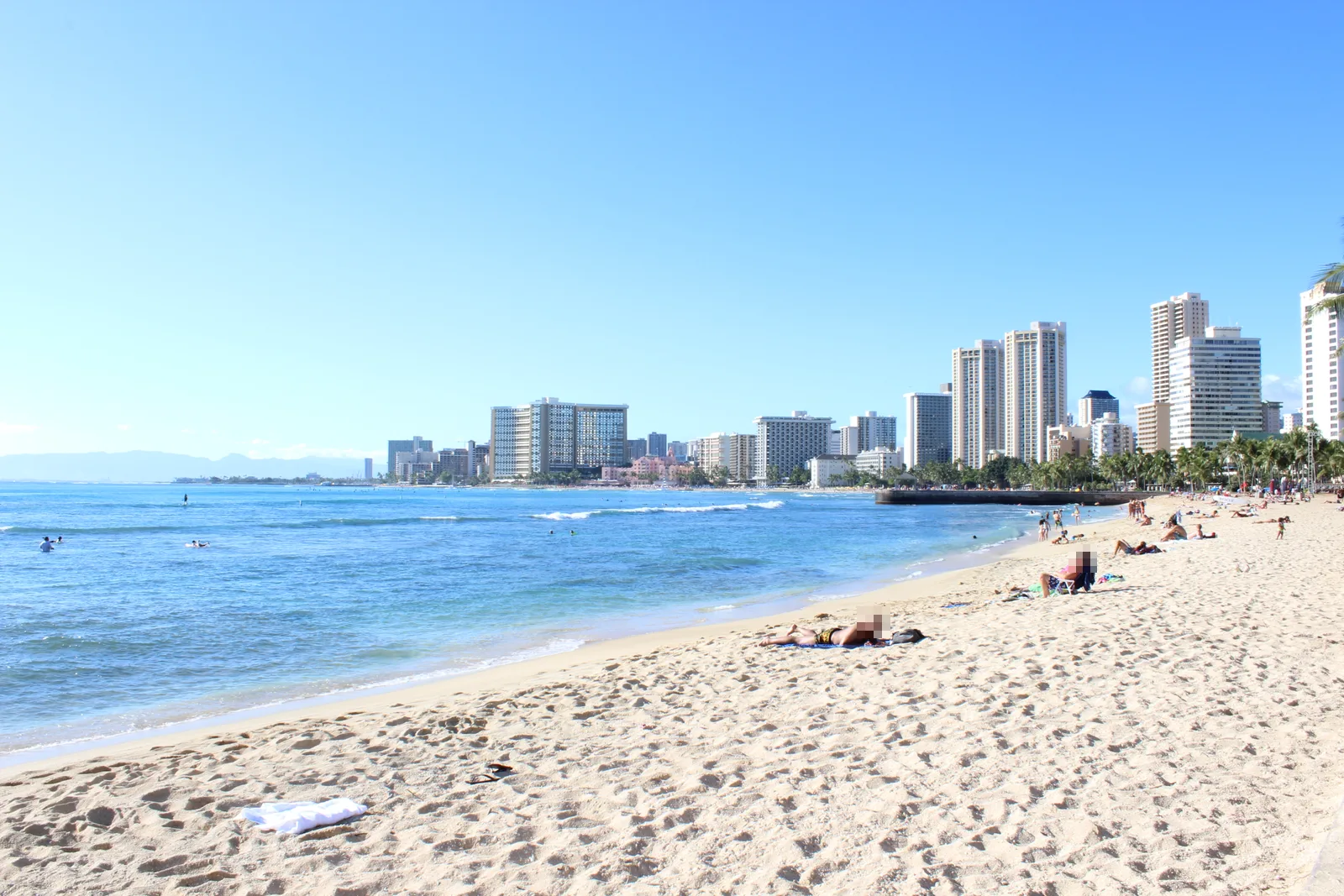 白い砂浜と青い海の向こうに、ワイキキのホテル群が弓なりに並ぶ。浜では数人が日光浴をしている
