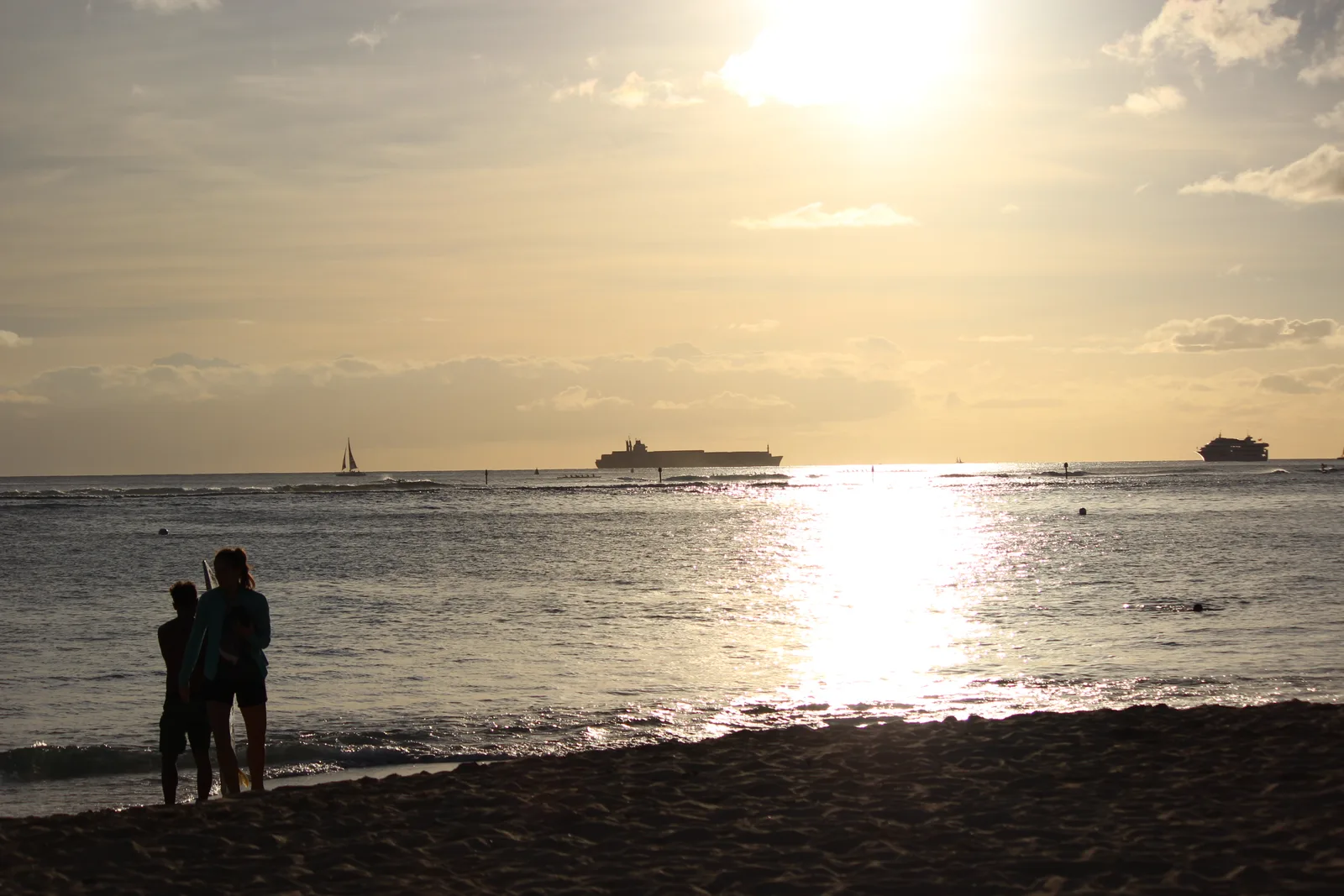 沖の船とともに金色に輝く夕方の海。浜辺に2人のシルエットが立っている