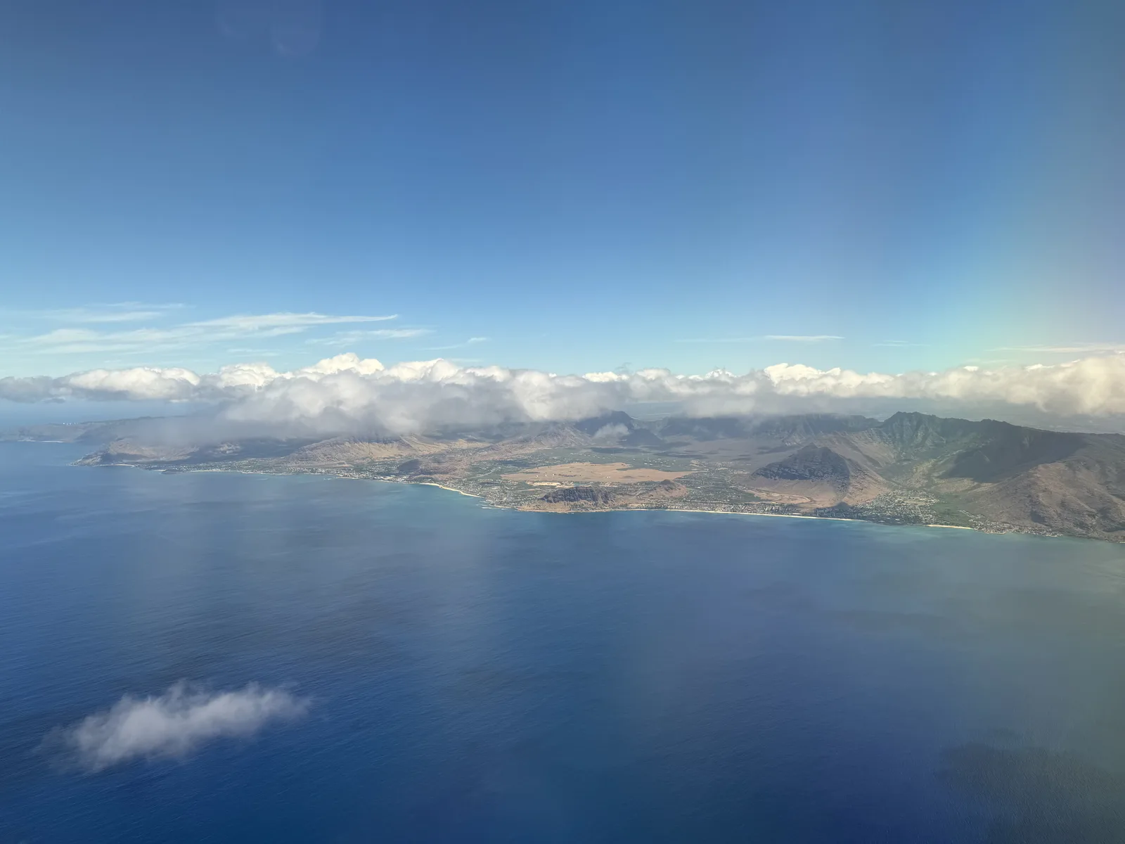 飛行機の窓から見た、青い海と雲のかかった山並み、海岸沿いに町が続くオアフ島。上には晴れた青空が広がっている