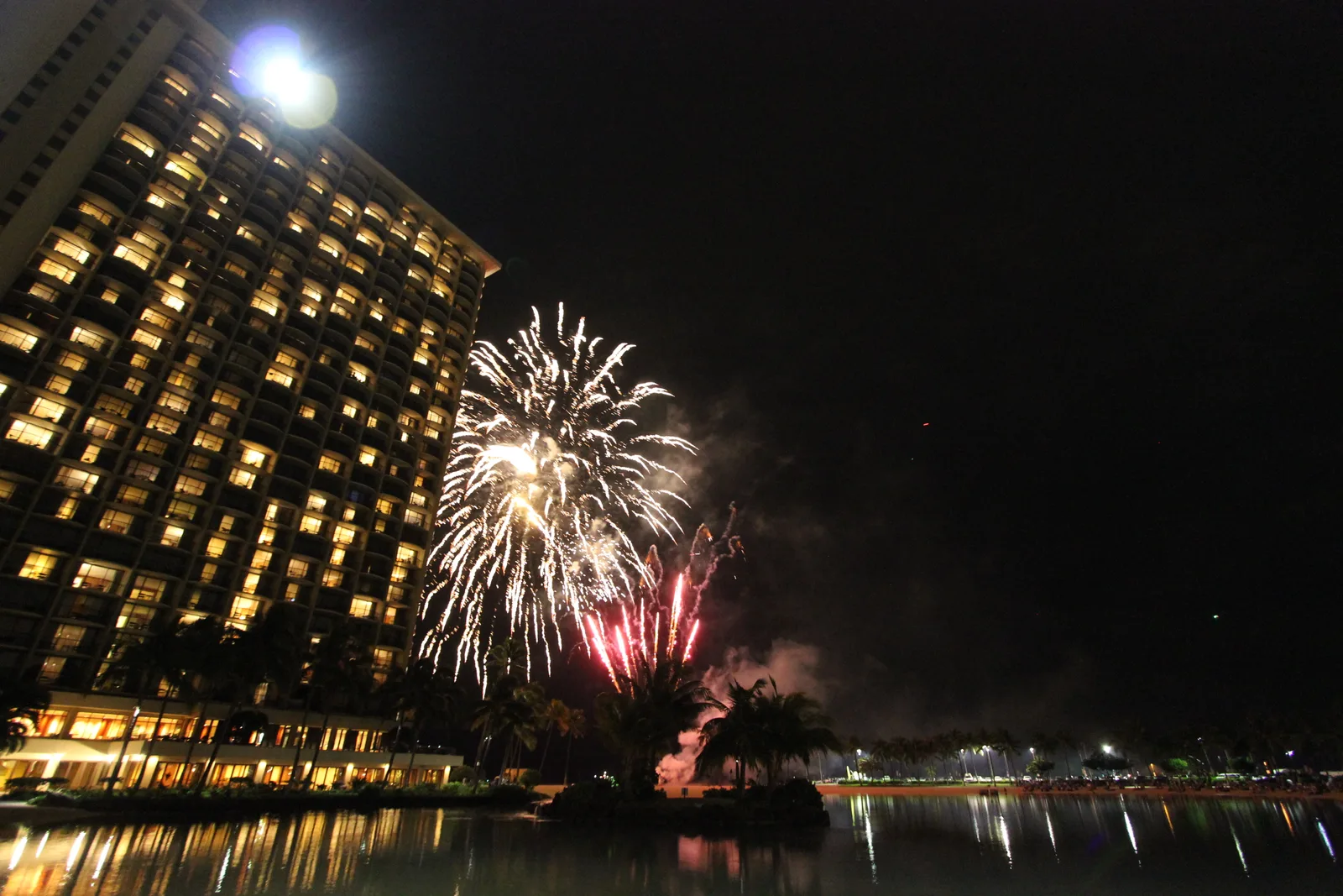 夜のラグーンの向こうで白い花火が開き、左に明かりの灯る高層タワーがそびえる。水面に光が映っている
