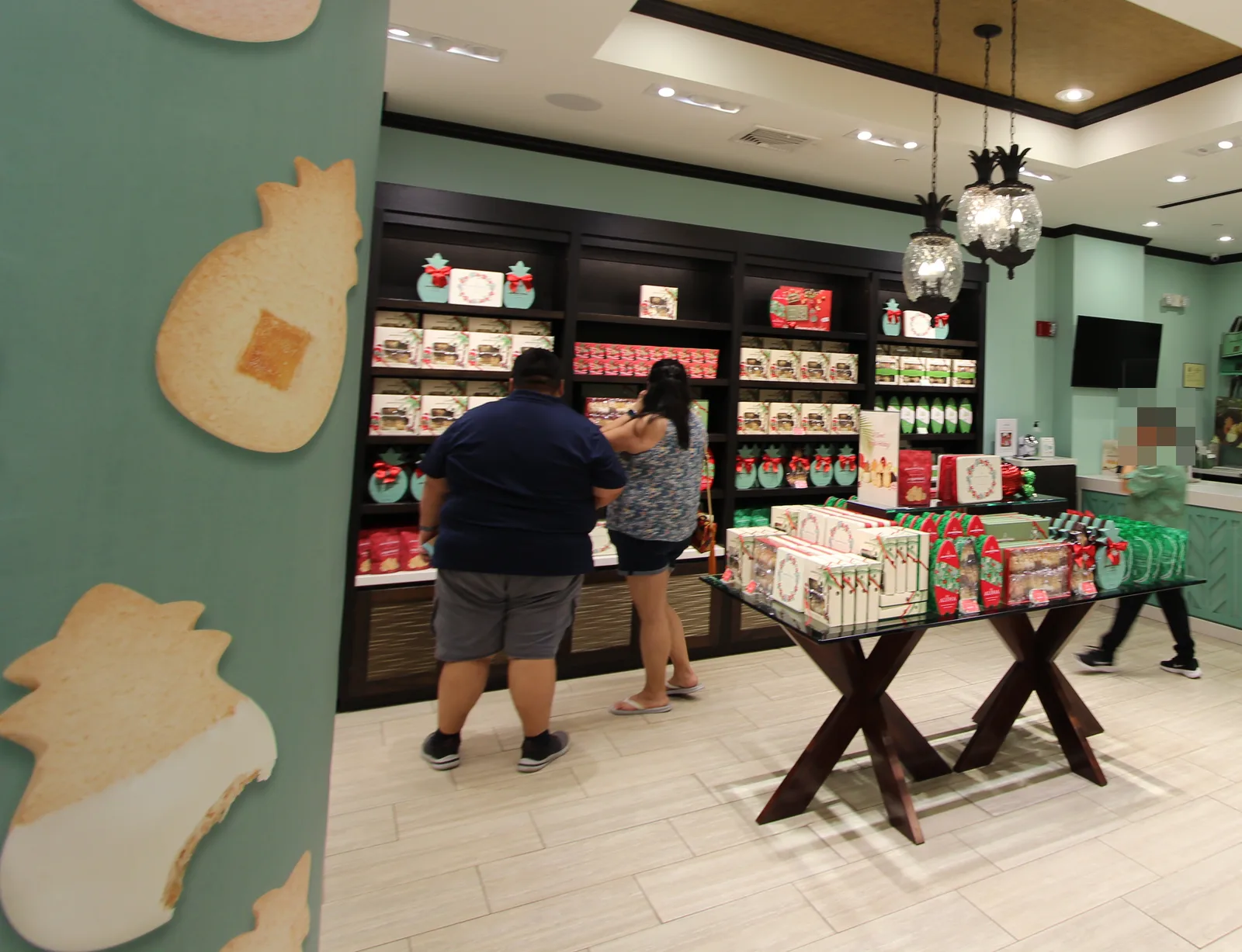 壁にパイナップル形のクッキーの絵が描かれた店内。奥の棚とテーブルに缶や箱が並び、買い物客が商品を選んでいる