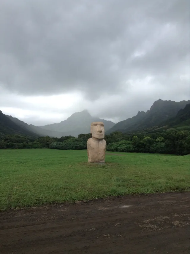 雲のかかった山並みを背に、草原にモアイ像のレプリカが1体立っている。手前は土の道