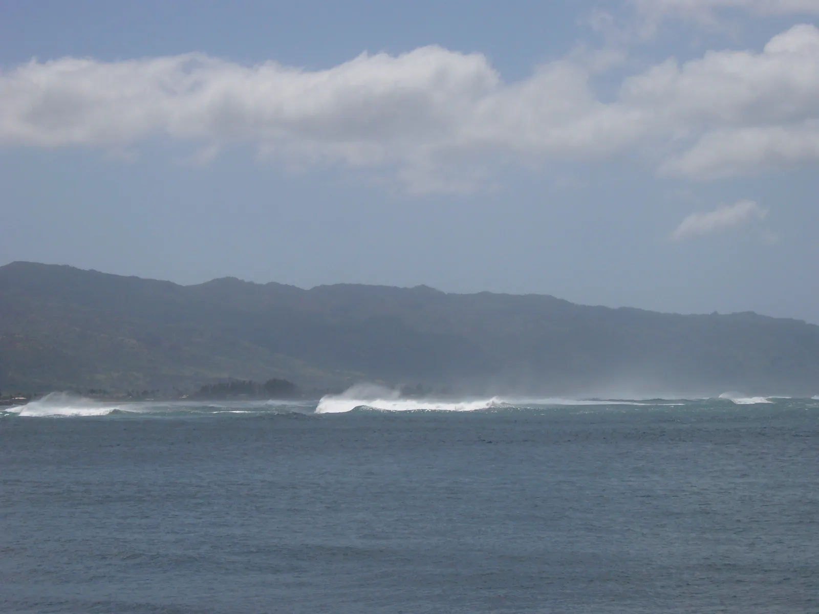 青い海の沖で白い波が砕けてしぶきが上がり、その奥に低い山並みが続いている