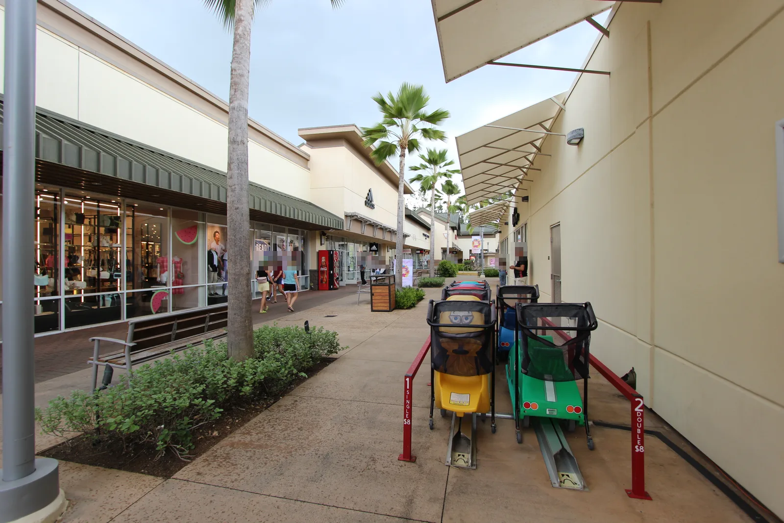 ヤシの木が並ぶ屋外の通路の両側に店舗が続き、手前に子ども用の車の形をしたカートが並んでいる