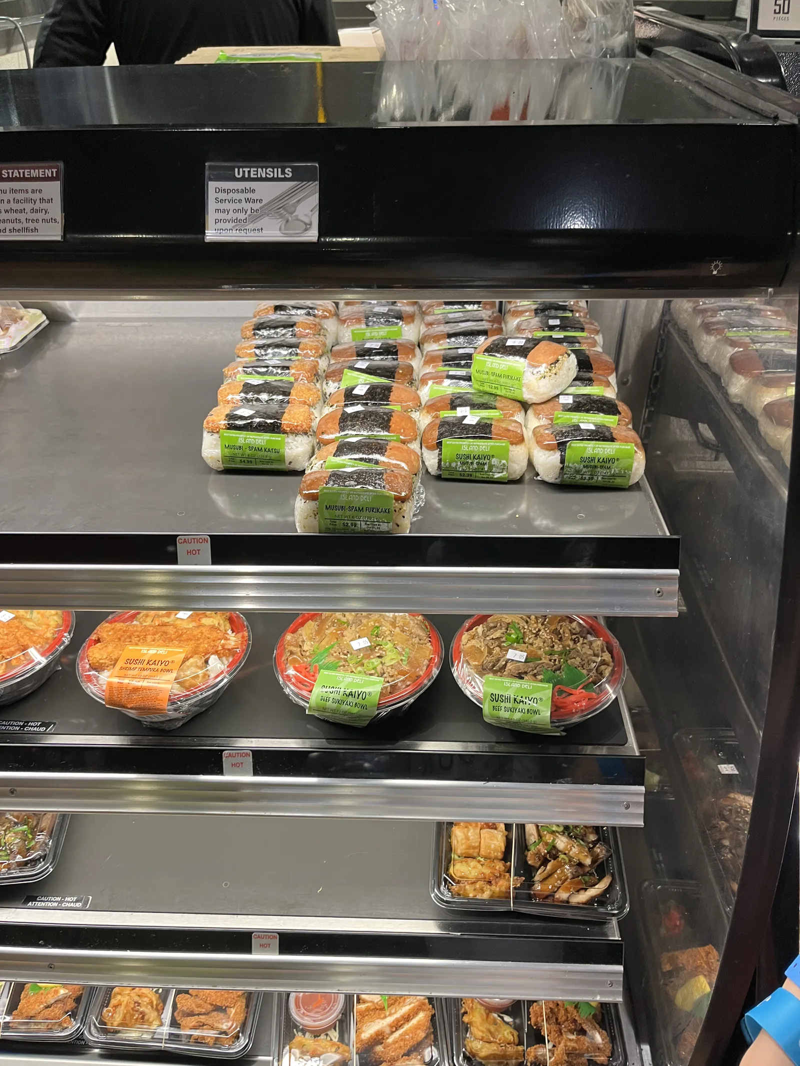 保温ケースに並んだ「ISLAND DELI」のラベルのスパムむすびと、天ぷらやすき焼きの丼