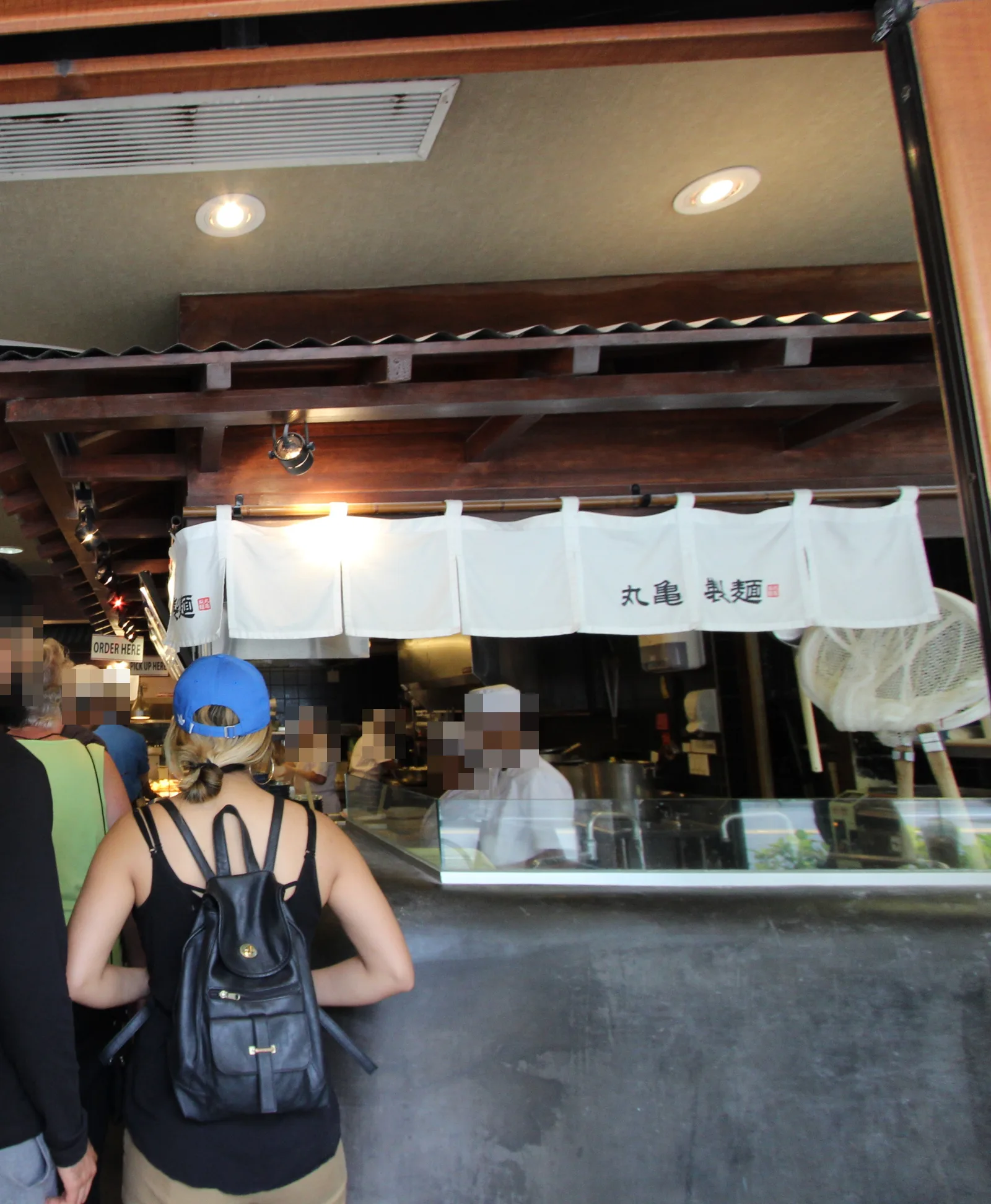 「丸亀製麺」と書かれた白いのれんの下で、白い帽子の店員がうどんを作るカウンターと、並んで待つお客さん
