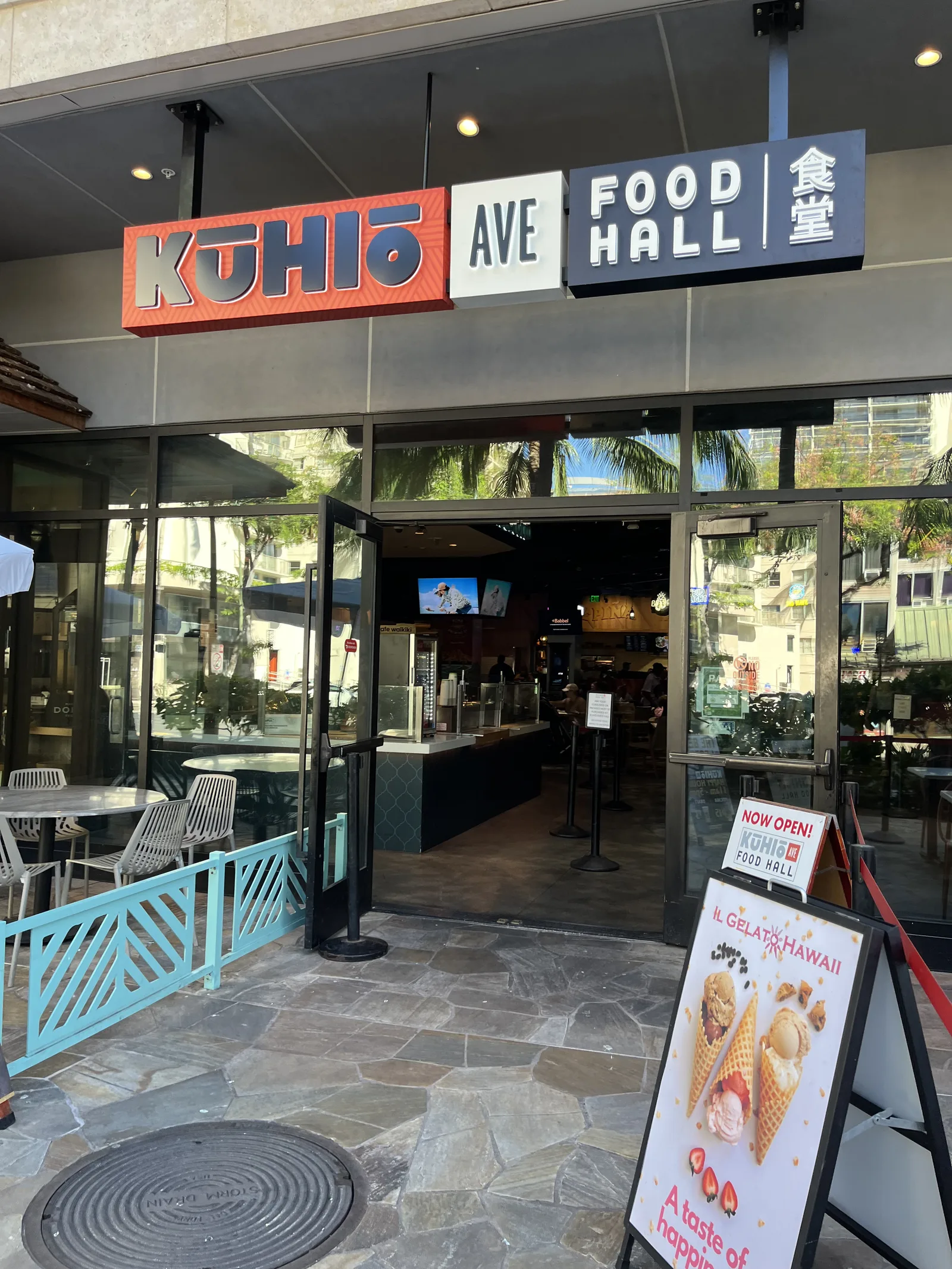 「KUHIO AVE FOOD HALL 食堂」の看板が掲げられたガラス張りの入口と、水色の柵で囲まれたテラス席