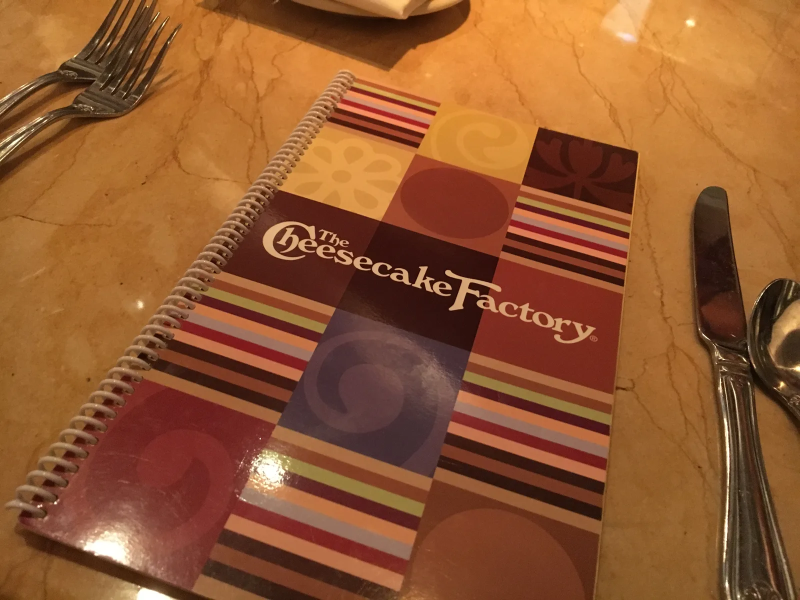 大理石のテーブルの上に置かれた、カラフルな表紙の「The Cheesecake Factory」の分厚いメニューとナイフ・フォーク